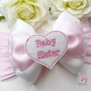 May include: A hair bow featuring white and light pink satin ribbons accented with small white beads. Centered on the bow is a white glittery heart-shaped patch with the pink embroidered text Baby Sister. The design is layered and symmetrical.
