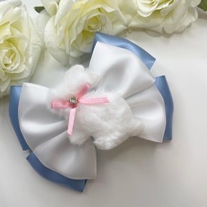 May include: A white and blue satin bow with a white fluffy bunny charm with a pink ribbon and a rhinestone in the centre.