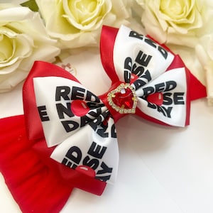 May include: Red nose day bow with a red heart-shaped rhinestone. The bow is made of white satin with black text that says "Red Nose Day" and red satin with white text that says "Red Nose Day".
