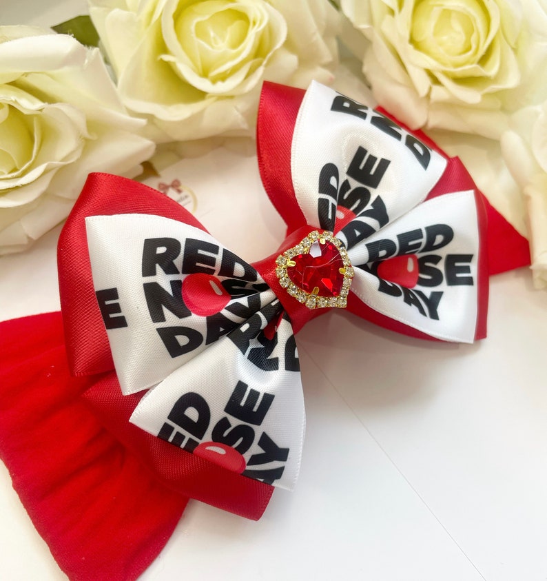 May include: Red nose day bow with a red heart-shaped rhinestone. The bow is made of white satin with black text that says "Red Nose Day" and red satin with white text that says "Red Nose Day".