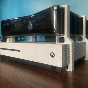 Puede incluir: Una consola Xbox 360 negra se encuentra encima de una consola Xbox One S blanca, sostenida por un soporte blanco impreso en 3D. La Xbox 360 es negra brillante con una bandeja de disco plateada. La Xbox One S tiene el logo de Xbox.