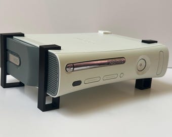 Op de afbeelding: Een off-white Xbox 360-console met een zwarte standaard. De console heeft een zilveren schijflade en een aan/uit-knop. De standaard is gemaakt van zwart plastic en verhoogt de console. Het is een gaming-apparaat.