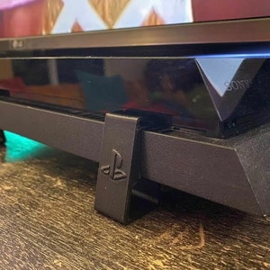 May include: Black stand with a PlayStation logo on the side, supporting a black Sony PlayStation console.