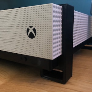 Pode incluir: Um console Xbox Series X branco com um logotipo Xbox preto na frente, sentado em um suporte preto com uma superfície texturizada. O console está em uma superfície de madeira.