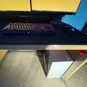 May include: A white computer tower sits under a desk with a black keyboard and mouse on a black mousepad. The desk has a wooden top and a wooden frame.