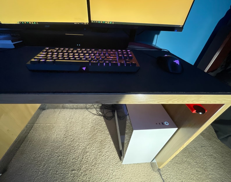 May include: A white computer tower sits under a desk with a black keyboard and mouse on a black mousepad. The desk has a wooden top and a wooden frame.