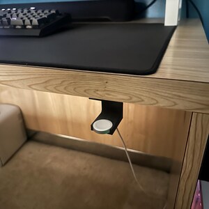Op de afbeelding: Een zwarte Apple Watch oplaadstandaard bevestigd onder een houten bureau. De standaard houdt een witte Apple Watch met een witte oplaadkabel vast. Een zwart toetsenbord en muismat zijn zichtbaar op het bureaublad.