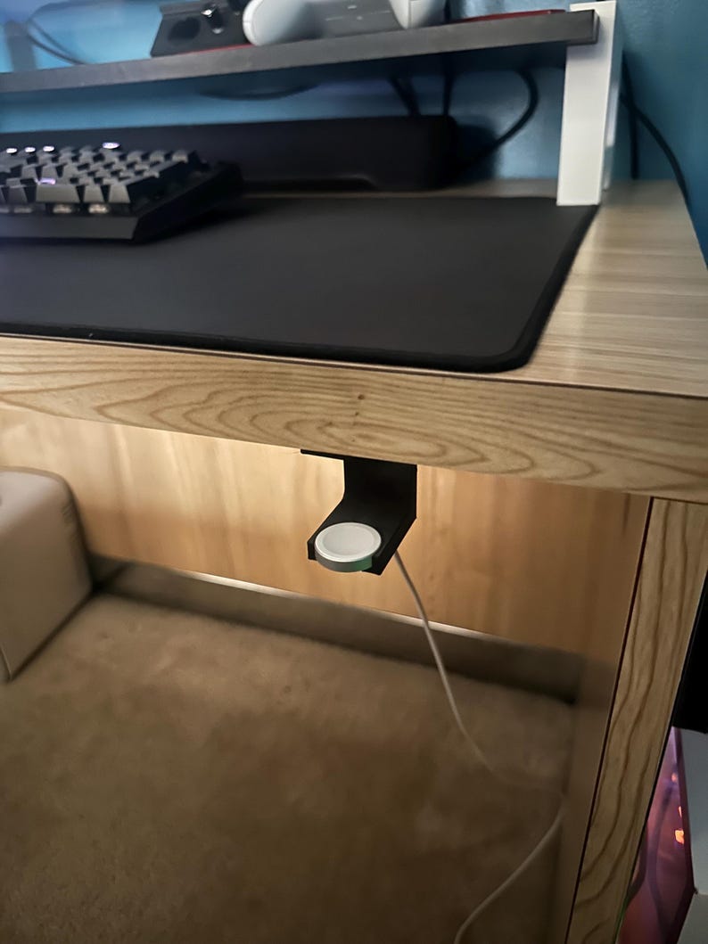 Op de afbeelding: Een zwarte Apple Watch oplaadstandaard bevestigd onder een houten bureau. De standaard houdt een witte Apple Watch met een witte oplaadkabel vast. Een zwart toetsenbord en muismat zijn zichtbaar op het bureaublad.