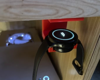 Op de afbeelding: Een zwarte smartwatch met een wit digitaal display dat "100%" laat zien, is gemonteerd op een rode 3D-geprinte houder. Het horloge is bevestigd aan een zwarte polsband.