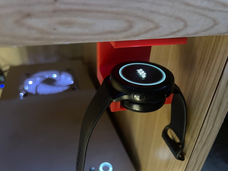 May include: A black smartwatch with a white digital display showing "100%" is mounted on a red 3D printed holder. The watch is attached to a black wristband.