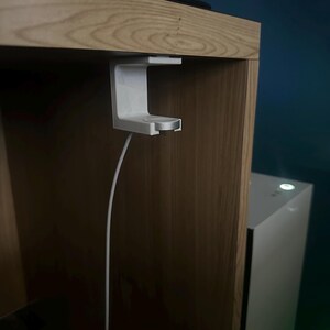Op de afbeelding: Een witte oplaadhouder voor een smartwatch, gemonteerd onder een houten plank. De houder heeft een cirkelvormig oplaadgedeelte en een witte kabel. Een computerkast is zichtbaar op de achtergrond.
