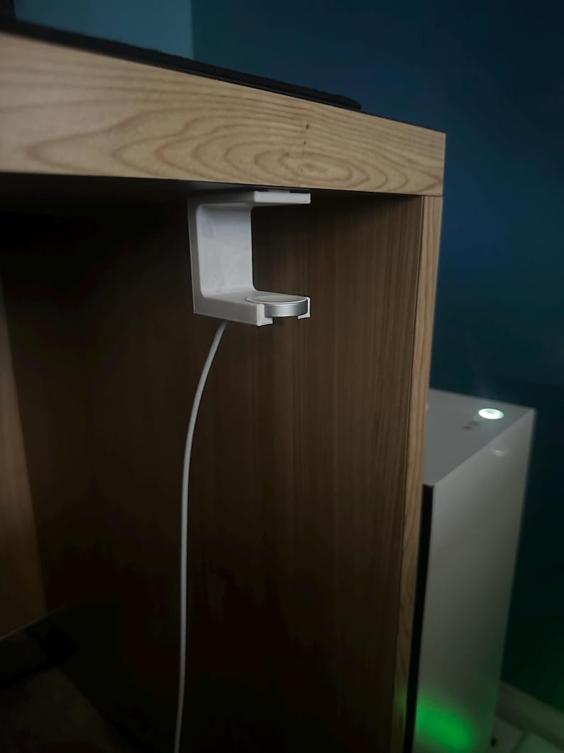 Op de afbeelding: Een witte oplaadhouder voor een smartwatch, gemonteerd onder een houten plank. De houder heeft een cirkelvormig oplaadgedeelte en een witte kabel. Een computerkast is zichtbaar op de achtergrond.