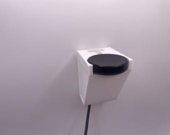 Op de afbeelding: Een witte wandbeugel met een zwart rond apparaat dat eraan is bevestigd. Het apparaat is aangesloten en heeft een snoer dat naar beneden hangt.