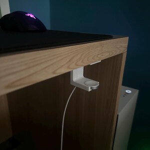 Op de afbeelding: Een witte Apple Watch oplaadstandaard is bevestigd aan de rand van een houten bureau. Een zwarte muis en muismat liggen op het bureau. Een witte oplaadkabel hangt naar beneden. De achtergrond is een donkerblauwe muur.