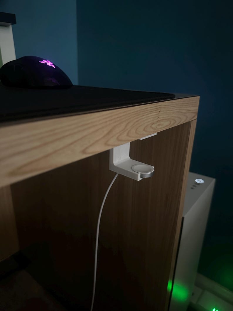 Op de afbeelding: Een witte Apple Watch oplaadstandaard is bevestigd aan de rand van een houten bureau. Een zwarte muis en muismat liggen op het bureau. Een witte oplaadkabel hangt naar beneden. De achtergrond is een donkerblauwe muur.