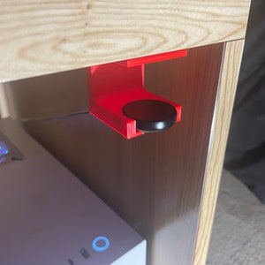May include: A red 3D printed hook mounted under a wooden shelf with a black round object hanging from it.