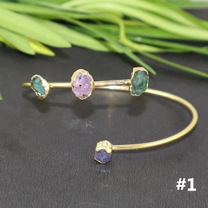 May include: A gold-toned open cuff bracelet featuring four raw, irregularly shaped gemstones. The stones include two green, one pink, and one blue crystal, each set in a gold-colored metal frame. The number #1 is printed in white in the corner.