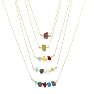 May include: Several gold-tone necklaces, each with a row of colourful, raw gemstones. The gemstones are in shades of red, green, yellow, blue, and brown. The necklaces are shown against a white backdrop.