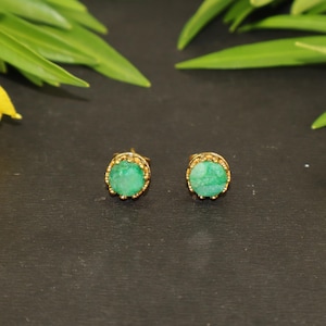Puede incluir: Un par de pendientes de botón con piedras preciosas verdes redondas y de corte rugoso, engastadas en monturas de metal dorado con un detalle de borde perlado. Las piedras tienen un aspecto texturizado y natural dentro de los marcos circulares.