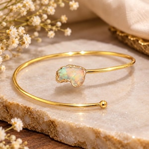 Peut inclure: Un bracelet ouvert doré doté d'une pierre brute aux reflets irisés à une extrémité et d'une petite sphère dorée à l'autre. La tige métallique est fine et polie, présentant un design délicat qui met en valeur la texture naturelle de la pierre.