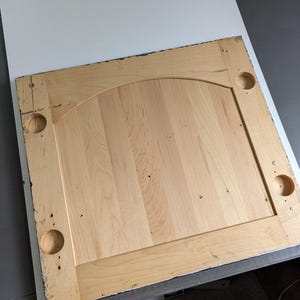 May include: A light-coloured wooden cabinet door with a raised panel and arched top. The door has four circular holes and is made of vertical wooden planks. The edges are framed with a darker trim.