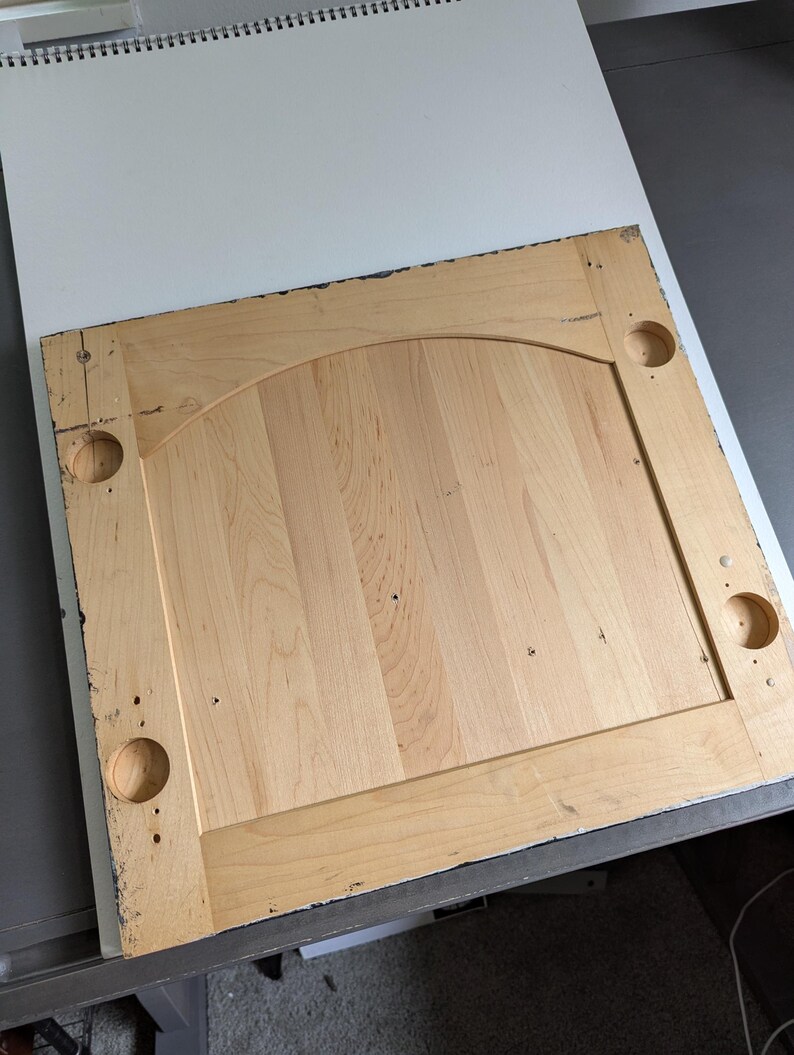 May include: A light-coloured wooden cabinet door with a raised panel and arched top. The door has four circular holes and is made of vertical wooden planks. The edges are framed with a darker trim.