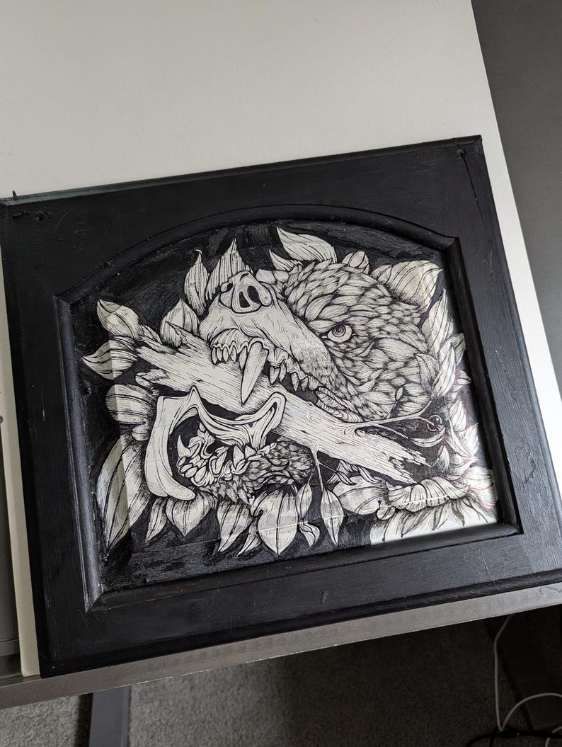 May include: Black framed artwork featuring a detailed pen and ink illustration of a wolf's head with a bone in its mouth, surrounded by leaves. The artwork is in a black frame.