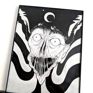 May include: Framed black and white artwork depicting a creature with large eyes, a wide mouth, and a crescent moon. The artwork is in a black frame with a black and white striped background. The artwork is in a black frame.