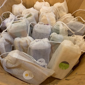 May include: A collection of handmade soaps in white mesh bags. The soaps are in various colors and scents, including "Forest Lemon" and "Basil & Mint".