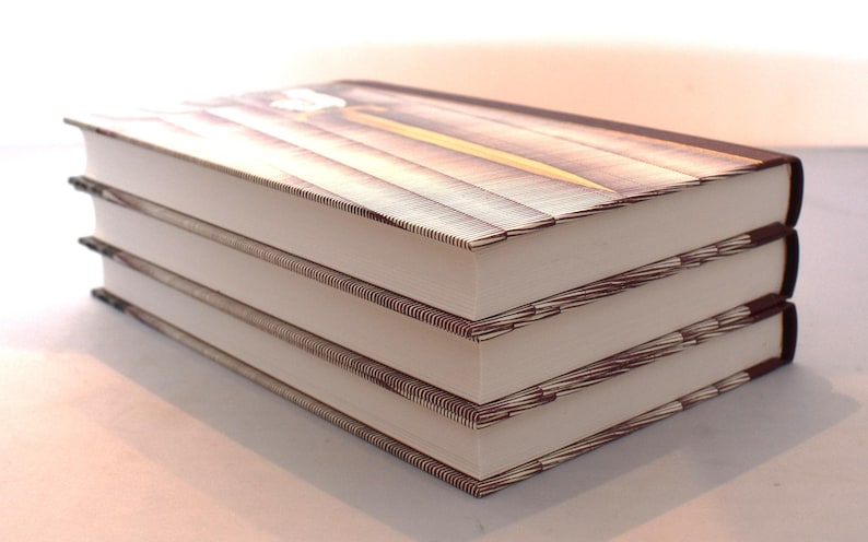 May include: A stack of three hardcover books with white pages and brown covers. The covers feature a wooden plank design with a yellow accent. The books are neatly stacked, suggesting a collection or set.