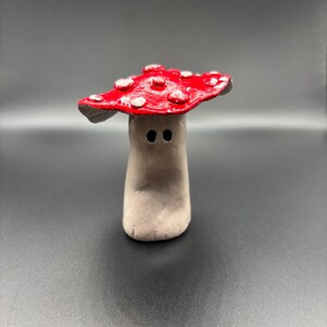 Könnte beinhalten: Eine kleine, handgeformte Tonfigur in Form eines Pilzes. Der Hut ist leuchtend rot mit erhabenen weißen Punkten bemalt, während der Stiel eine neutrale beigefarbene Tönung aufweist und zwei kleine, dunkle, kreisförmige Vertiefungen als Augen hat.