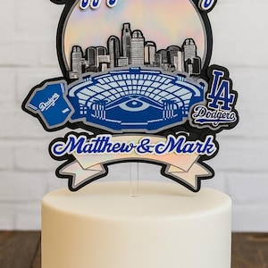 May include: A white cake with a blue ribbon base, topped with a Dodgers-themed cake topper. The topper features a baseball, the words "Happy Birthday", a city skyline, a baseball stadium, and the names "Matthew & Mark".