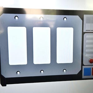 May include: A gray and black wall plate with three rectangular openings. The plate has a control panel design on the right side with a red button, blue and red squares, and a series of gray rectangles.