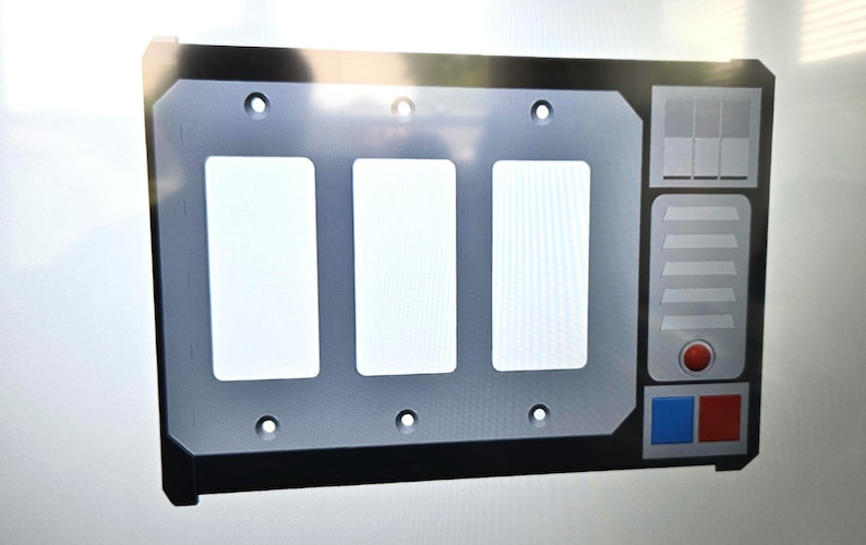 May include: A gray and black wall plate with three rectangular openings. The plate has a control panel design on the right side with a red button, blue and red squares, and a series of gray rectangles.