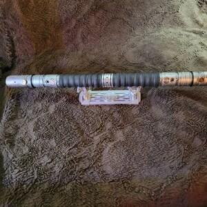 May include: A silver lightsaber with a black grip and a silver stand. The lightsaber is on a brown fuzzy surface.