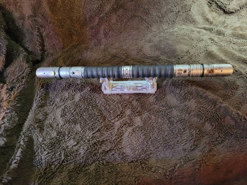 May include: A silver lightsaber with a black grip and a silver stand. The lightsaber is on a brown fuzzy surface.