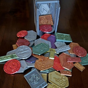 May include: A collection of colorful plastic tokens with various symbols and text, including "Dice 10", "Command", and "Force", scattered on a wooden surface. A gray plastic box with a grid pattern and the number "4" on the front is tipped over, spilling the tokens.