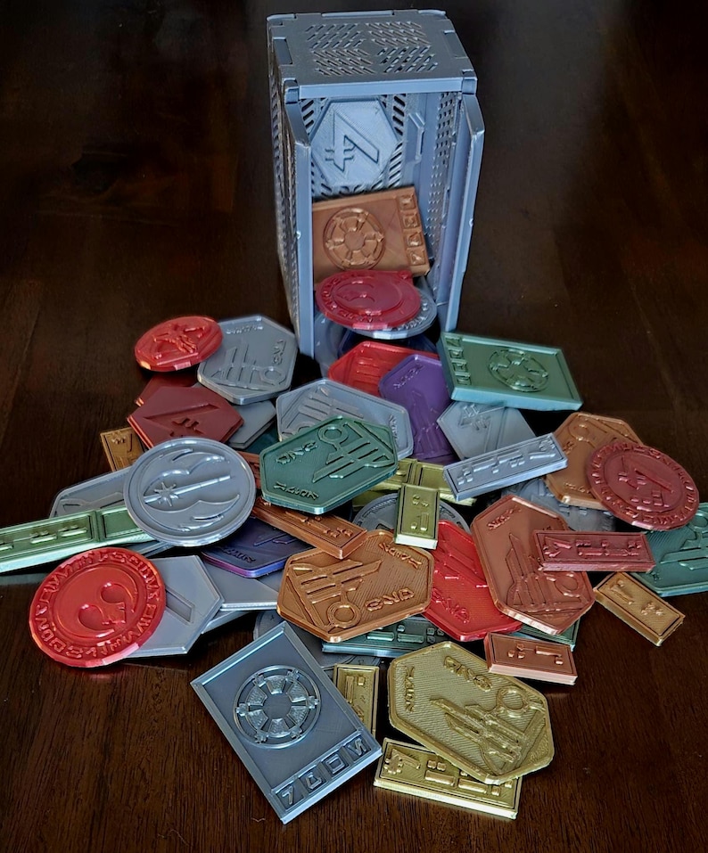 May include: A collection of colorful plastic tokens with various symbols and text, including "Dice 10", "Command", and "Force", scattered on a wooden surface. A gray plastic box with a grid pattern and the number "4" on the front is tipped over, spilling the tokens.