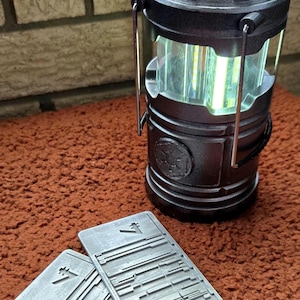 Peut inclure: Une lanterne cylindrique noire avec une source de lumi&egrave;re verticale claire. Deux objets rectangulaires argent&eacute;s avec des motifs grav&eacute;s sont au premier plan. La lanterne a une poign&eacute;e et repose sur une surface marron textur&eacute;e.