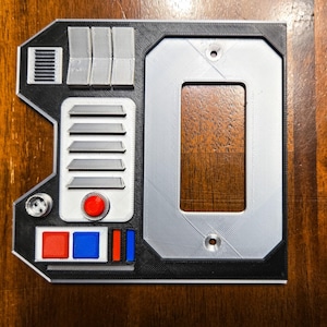 May include: A black and gray 3D printed wall plate with a rectangular cutout, a red button, and a series of gray lines and rectangles.