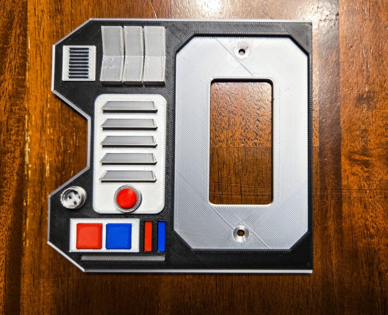 May include: A black and gray 3D printed wall plate with a rectangular cutout, a red button, and a series of gray lines and rectangles.