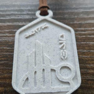 Puede incluir: Colgante blanco con cord&oacute;n marr&oacute;n. El colgante tiene una forma geom&eacute;trica con un dise&ntilde;o de paisaje urbano estilizado y la palabra "ONE" en una fuente &uacute;nica. Tambi&eacute;n es visible la palabra "2017".