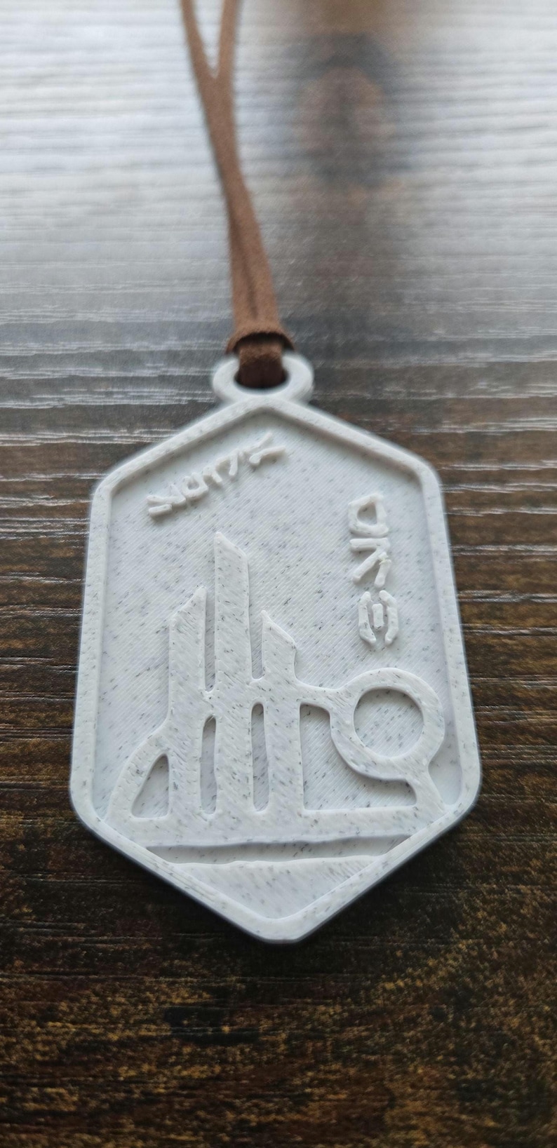 Puede incluir: Colgante blanco con cord&oacute;n marr&oacute;n. El colgante tiene una forma geom&eacute;trica con un dise&ntilde;o de paisaje urbano estilizado y la palabra "ONE" en una fuente &uacute;nica. Tambi&eacute;n es visible la palabra "2017".