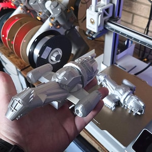 Puede incluir: Un modelo plateado impreso en 3D de una nave espacial se sostiene en una mano. El modelo tiene alas detalladas, una cabina y componentes del motor. Un segundo modelo similar está sobre una cama de impresora. Una impresora 3D y bobinas de filamento son visibles en el fondo.