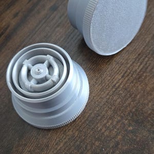 Puede incluir: Dos objetos grises impresos en 3D sobre una superficie de madera marr&oacute;n. Uno es de forma cil&iacute;ndrica con una parte superior texturizada. El otro es un dise&ntilde;o circular complejo con m&uacute;ltiples capas y una caracter&iacute;stica central.