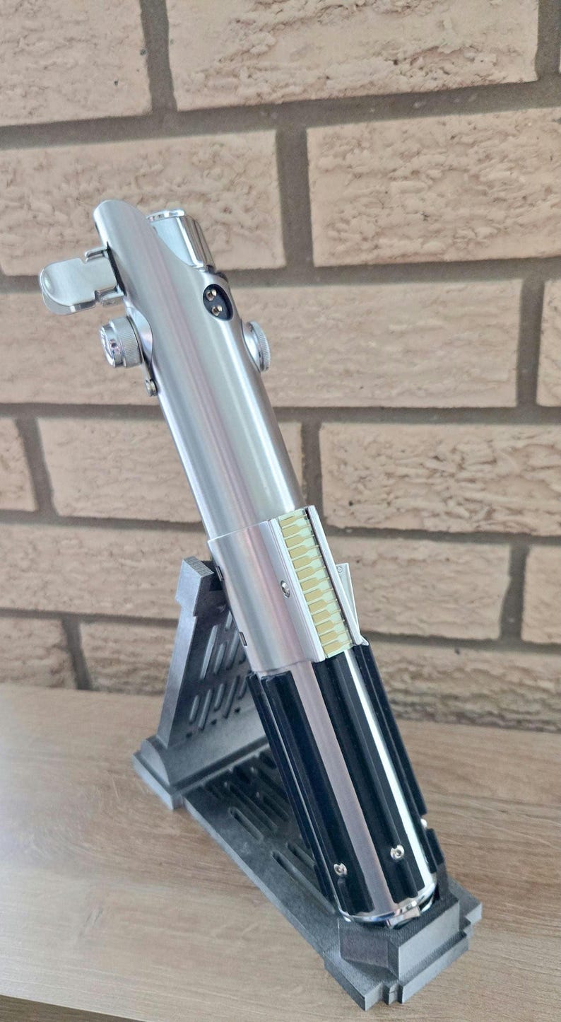 Op de afbeelding: Een zilveren lightsaber met zwarte accenten en een geel detail aan de zijkant, getoond op een zwarte standaard.