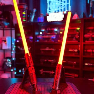 K&ouml;nnte beinhalten: Zwei rot-schwarze Lichtschwerter mit leuchtend gelben Klingen werden auf einer roten Matte pr&auml;sentiert. Die Lichtschwerter sind beleuchtet, und der Hintergrund zeigt ein Star Wars-Schild und andere Sammlerst&uuml;cke.