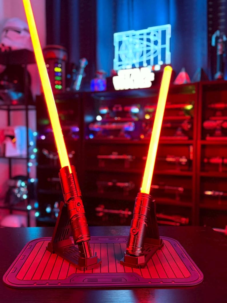 K&ouml;nnte beinhalten: Zwei rot-schwarze Lichtschwerter mit leuchtend gelben Klingen werden auf einer roten Matte pr&auml;sentiert. Die Lichtschwerter sind beleuchtet, und der Hintergrund zeigt ein Star Wars-Schild und andere Sammlerst&uuml;cke.