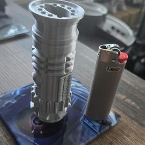 May include: A 3D printed model of a lightsaber, approximately 6 inches tall, standing on a blue and black circular base. A silver lighter with a red top is next to the lightsaber for scale.