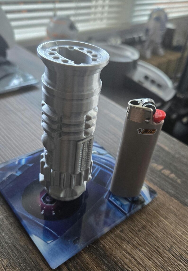 May include: A 3D printed model of a lightsaber, approximately 6 inches tall, standing on a blue and black circular base. A silver lighter with a red top is next to the lightsaber for scale.